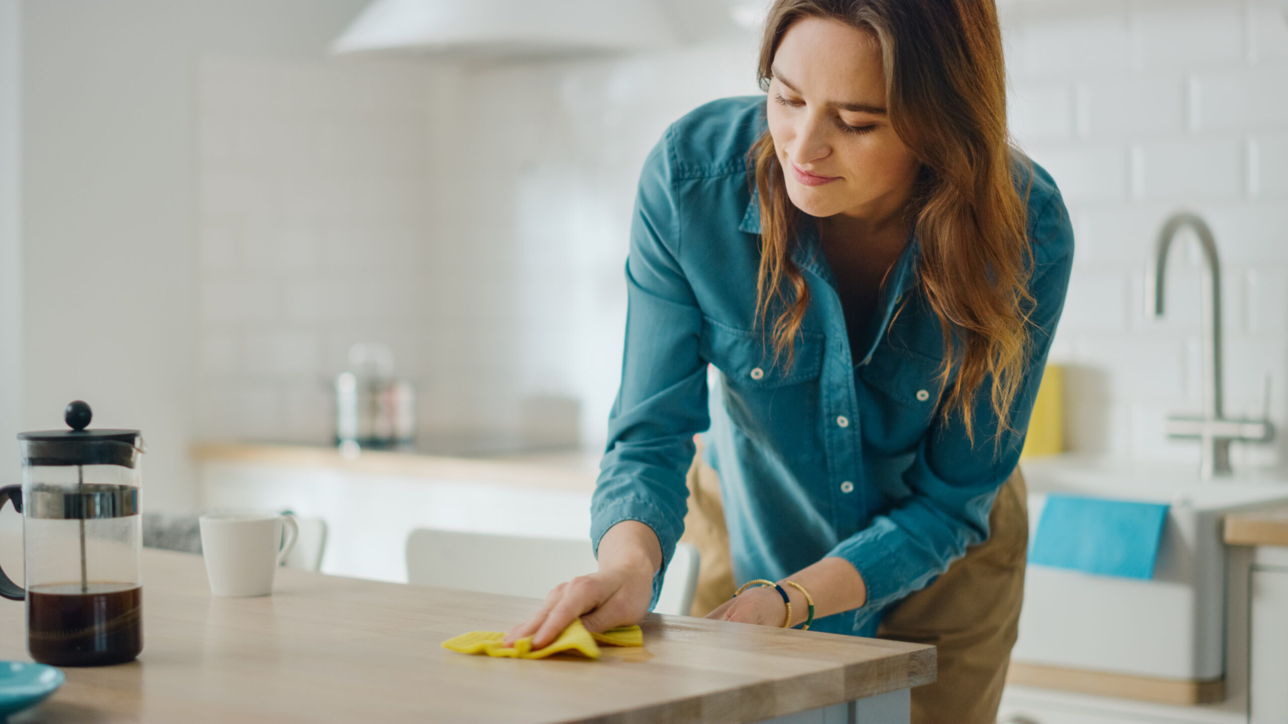 Caring Housewife Wiping Spilled Coffee or Crumbs from a Wooden Kitchen Table Beautiful Young Female Using a Household Cleaner Wipe for Cleaning Brunette is Wearing a Jeans Shirt and Beige Pants | MOLLY MAID