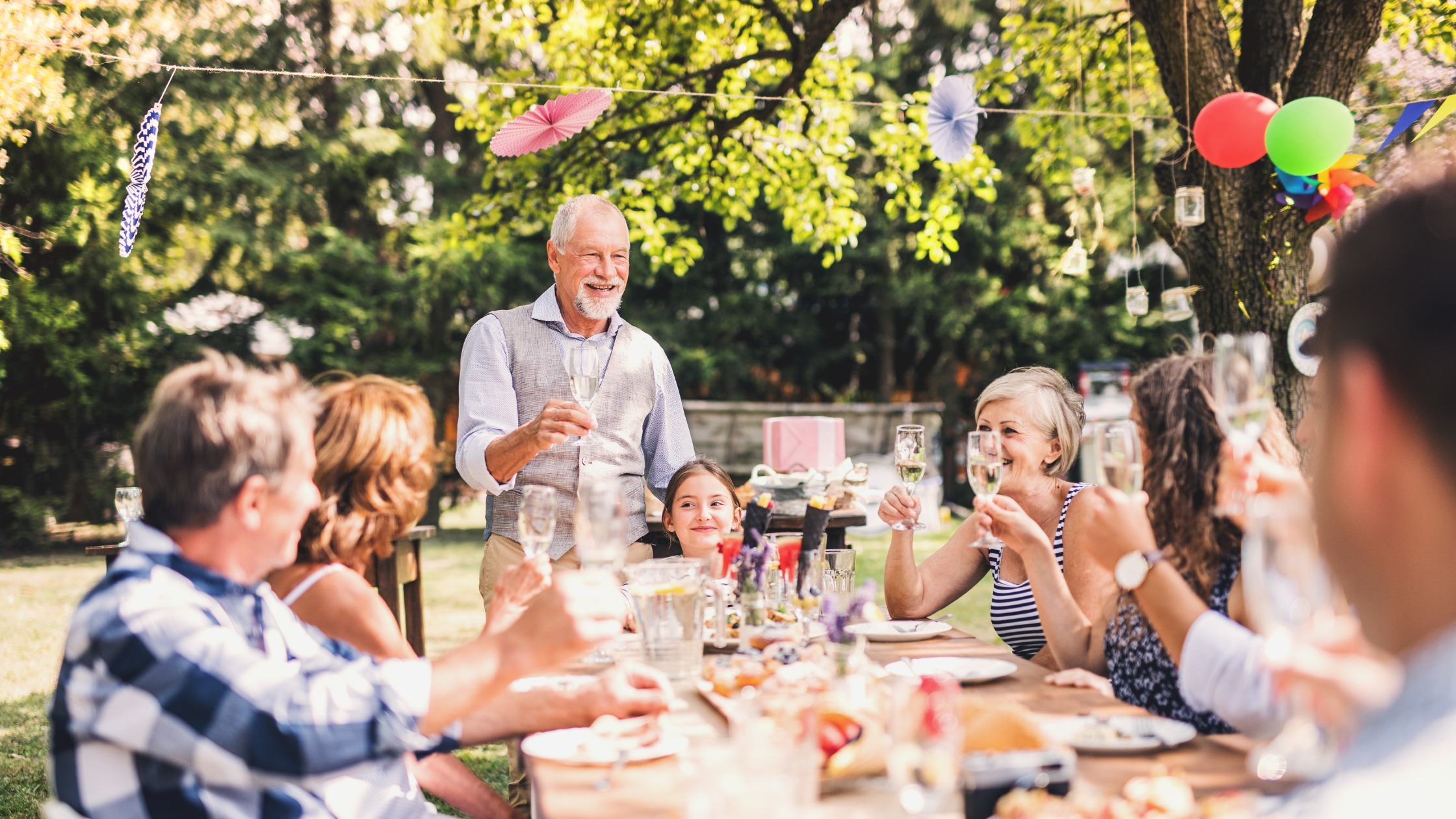 Group of adults and a child toasting with wine glasses at an outdoor garden party with colorful balloons and string lights overhead