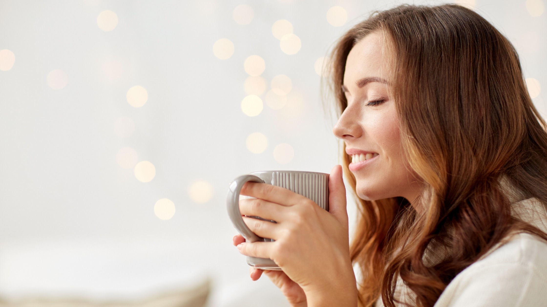 Woman with long brown hair smiling with eyes closed while sipping from a gray mug in a cozy softly lit setting with bokeh lights in the background