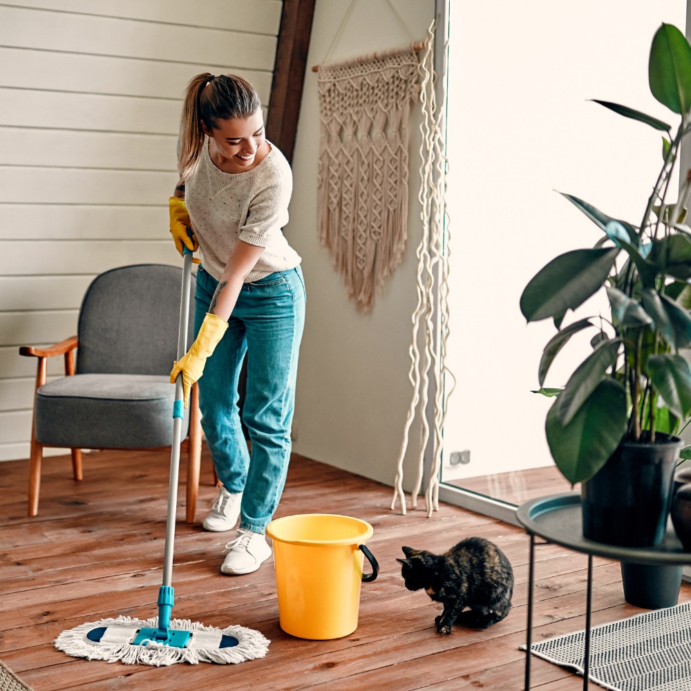 Woman mopping a wooden floor with a mop; wearing yellow gloves, white sweater, blue jeans, white sneakers, in a cozy room with a plant and cat nearby.