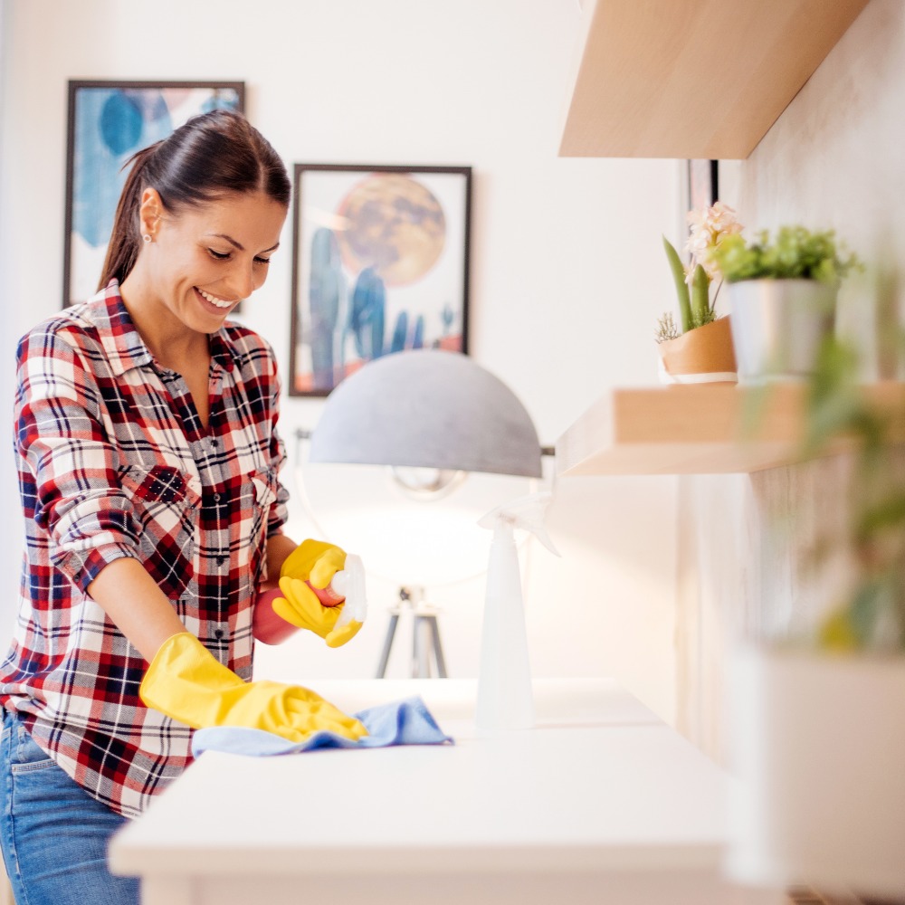 Woman wearing yellow cleaning gloves wipes a white countertop with a spray bottle and cloth, smiling in a bright room.