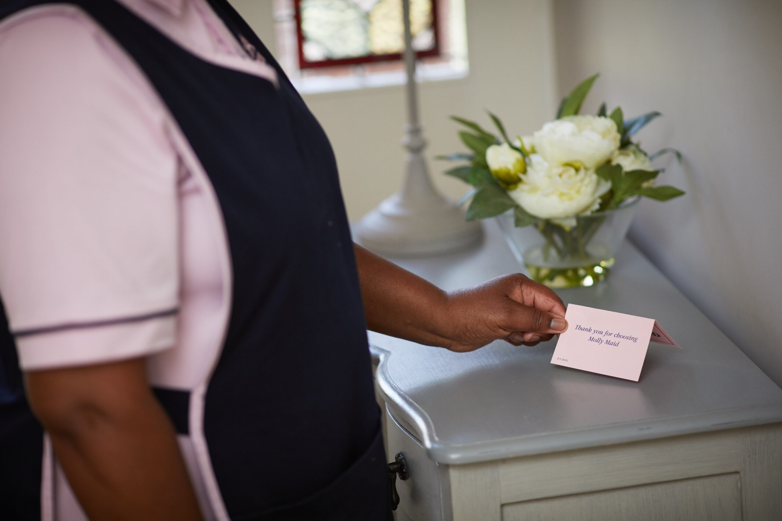 Person in a pink uniform and dark apron hands a pink Thank you card to someone at a counter with white flowers in the background