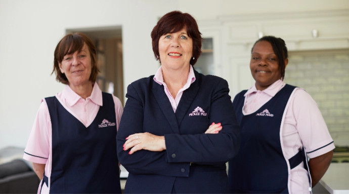 Three female staff members in pink polo shirts and navy vests pose together in a bright homey setting smiling at the camera
