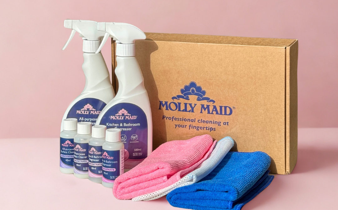 Cleaning supplies set with Molly Maid bottles a cardboard Molly Maid box and pink and blue microfiber cloths on a pink background